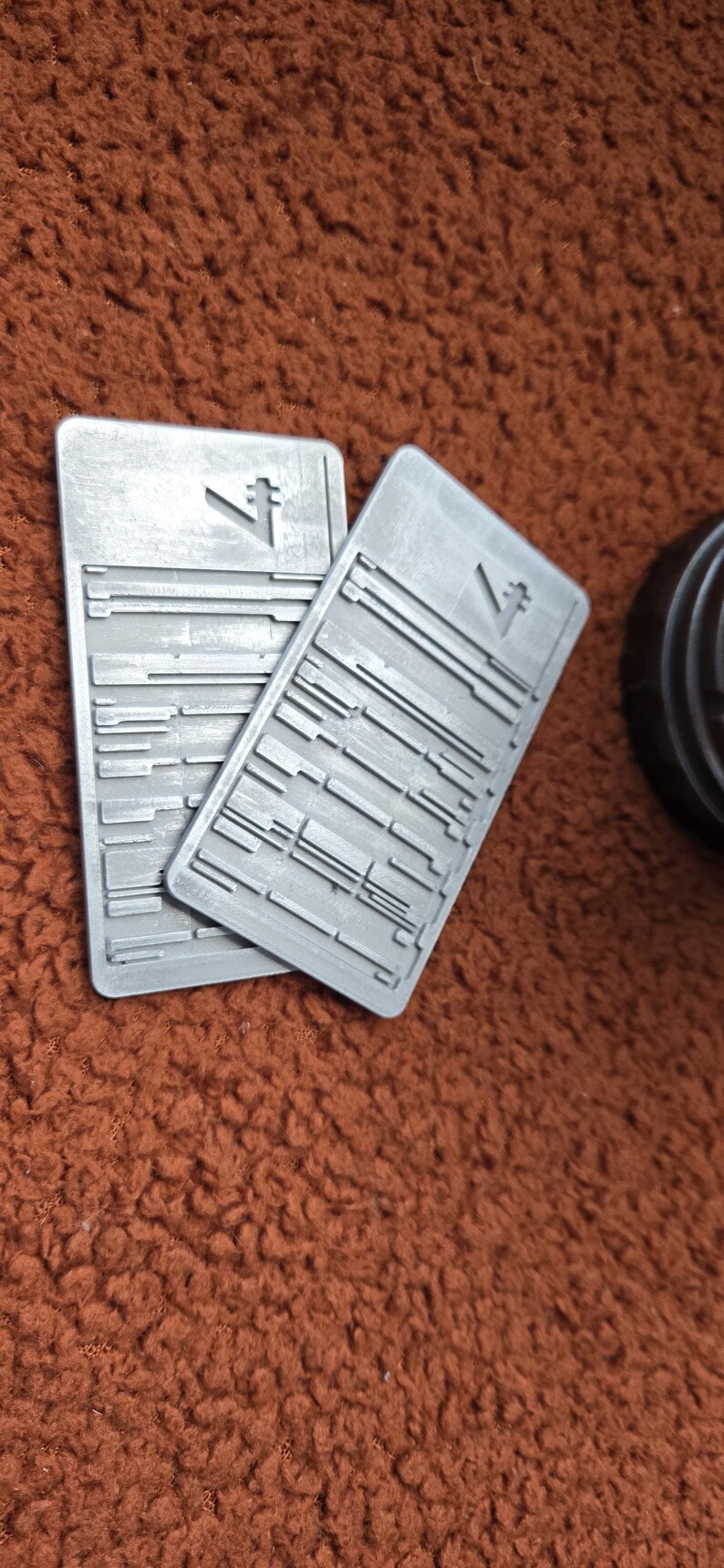Peut inclure: Deux objets rectangulaires gris avec une surface textur&eacute;e et un symbole stylis&eacute;. Les objets pr&eacute;sentent une s&eacute;rie de lignes horizontales et verticales. Les objets sont sur une surface marron textur&eacute;e.