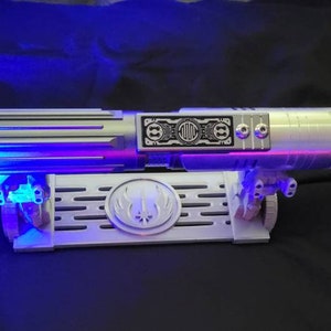 May include: A silver lightsaber with blue and purple lights on a white stand with a symbol on it. The lightsaber has a black panel with the text "D.O.C." on it.