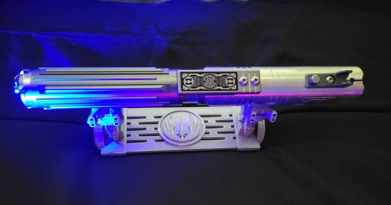 May include: A silver lightsaber with blue and purple lights on a white stand with a symbol on it. The lightsaber has a black panel with the text "D.O.C." on it.