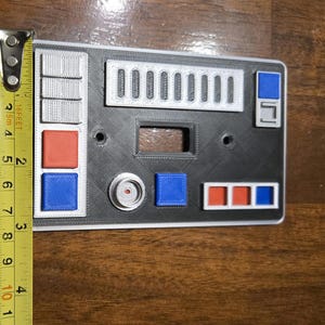 Op de afbeelding: Een zwart-witte wandplaat met verschillende knoppen en schakelaars. De plaat heeft een rood-blauw-wit kleurenschema. De plaat is 10 cm breed.