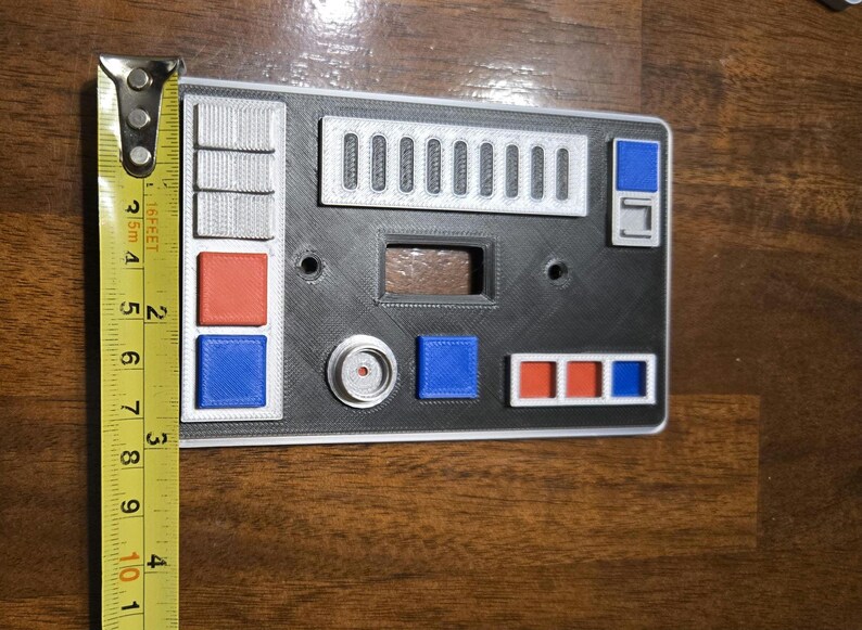 Op de afbeelding: Een zwart-witte wandplaat met verschillende knoppen en schakelaars. De plaat heeft een rood-blauw-wit kleurenschema. De plaat is 10 cm breed.