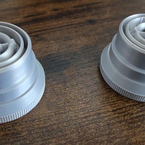 Puede incluir: Dos tapones de botella de pl&aacute;stico gris, con base estriada y parte superior circular detallada. Los tapones presentan anillos conc&eacute;ntricos y un dise&ntilde;o central en forma de estrella. Probablemente cierres de botellas o recipientes.
