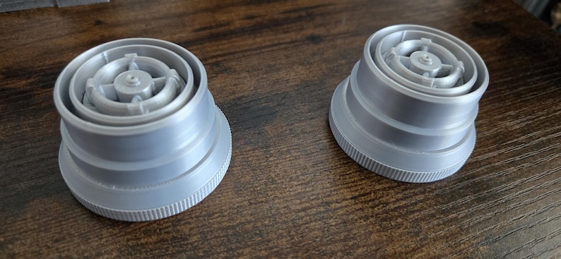 Puede incluir: Dos tapones de botella de pl&aacute;stico gris, con base estriada y parte superior circular detallada. Los tapones presentan anillos conc&eacute;ntricos y un dise&ntilde;o central en forma de estrella. Probablemente cierres de botellas o recipientes.