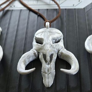 May include: Three white, textured, skull-shaped pendants with curved horns, each suspended from a brown cord. The pendants feature hollow eye sockets and a central nasal cavity, displaying a detailed, layered surface finish consistent with 3D printing.