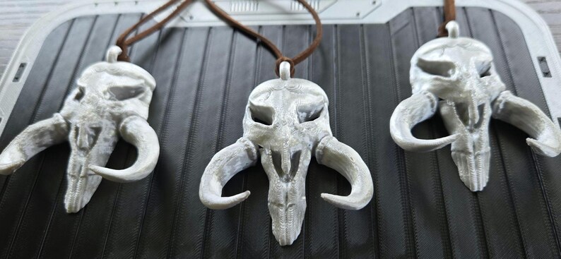 May include: Three white, textured, skull-shaped pendants with curved horns, each suspended from a brown cord. The pendants feature hollow eye sockets and a central nasal cavity, displaying a detailed, layered surface finish consistent with 3D printing.