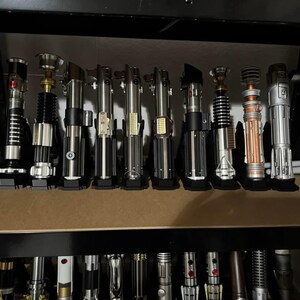 K&ouml;nnte beinhalten: Eine Sammlung von Lichtschwertern, die auf einem Regal ausgestellt sind. Die Lichtschwerter haben unterschiedliche Designs mit silbernen, schwarzen und bronzefarbenen Griffen. Einige haben rote oder blaue Aktivierungskn&ouml;pfe. Das Regal ist aus dunklem Holz.