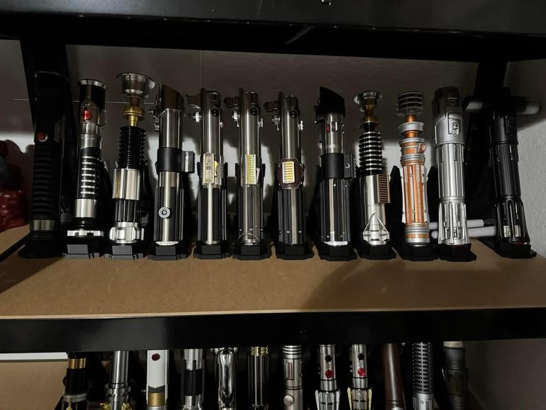K&ouml;nnte beinhalten: Eine Sammlung von Lichtschwertern, die auf einem Regal ausgestellt sind. Die Lichtschwerter haben unterschiedliche Designs mit silbernen, schwarzen und bronzefarbenen Griffen. Einige haben rote oder blaue Aktivierungskn&ouml;pfe. Das Regal ist aus dunklem Holz.