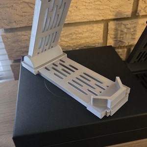 May include: A light gray 3D-printed stand with a rectangular base and an angled support. The support features vertical and horizontal slots. The stand is designed to hold an electronic device. The stand is on a black surface.