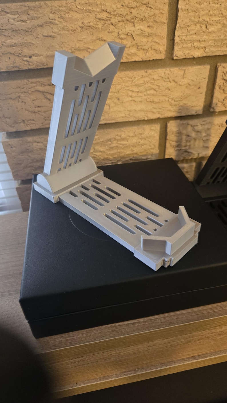 May include: A light gray 3D-printed stand with a rectangular base and an angled support. The support features vertical and horizontal slots. The stand is designed to hold an electronic device. The stand is on a black surface.