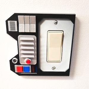 May include: A black and white light switch cover with a futuristic design. It features a light switch, a red button, and several other buttons and switches.