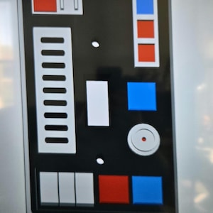 May include: A black control panel with a variety of buttons and switches. The panel has a white rectangular area with a series of small white rectangles, a white square, and a white circle with a red dot in the center. The panel also has a red button, a blue button, and a red and blue button.