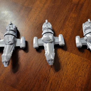 May include: Three silver-colored miniature spaceships, each with a distinctive design, are arranged in a row on a wooden surface.