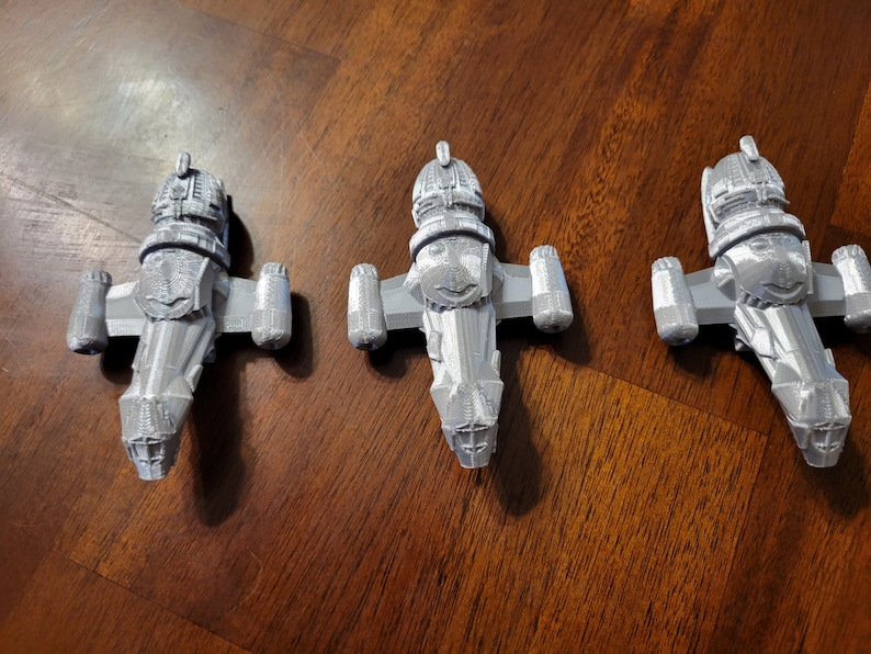 May include: Three silver-colored miniature spaceships, each with a distinctive design, are arranged in a row on a wooden surface.