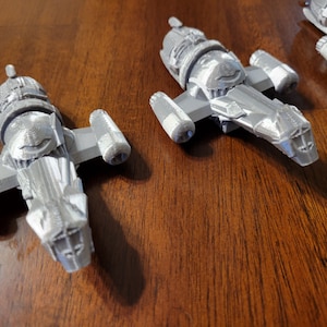 May include: Three gray, detailed, plastic model spaceships. The spaceships are all the same design, but are different sizes. The spaceships are sitting on a wooden surface.