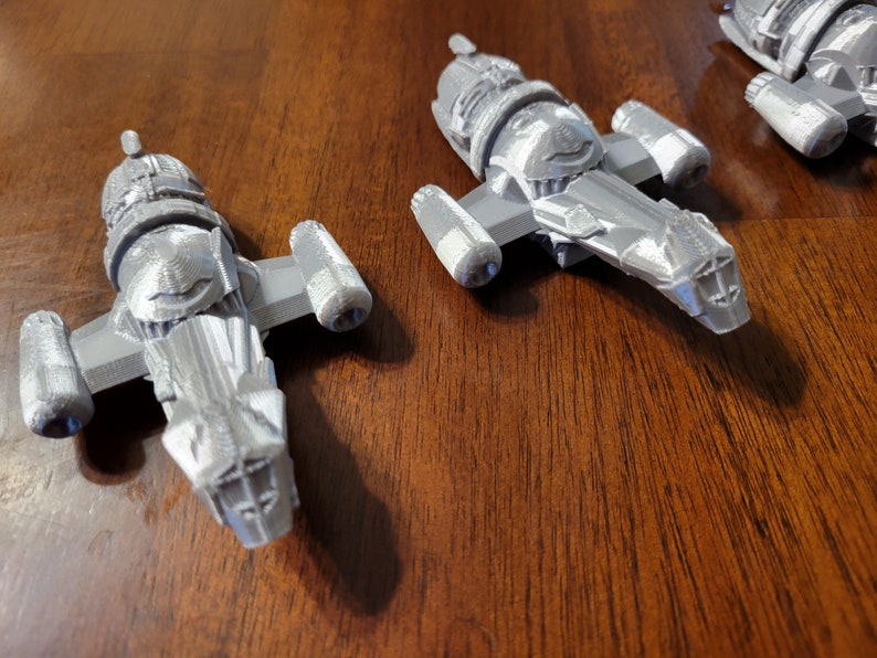 May include: Three gray, detailed, plastic model spaceships. The spaceships are all the same design, but are different sizes. The spaceships are sitting on a wooden surface.