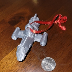 May include: A silver 3D printed spaceship ornament with a red ribbon attached. The ornament is about the size of a US quarter.