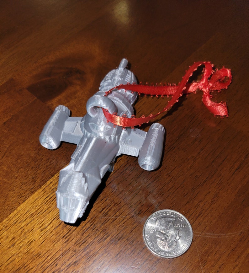 May include: A silver 3D printed spaceship ornament with a red ribbon attached. The ornament is about the size of a US quarter.