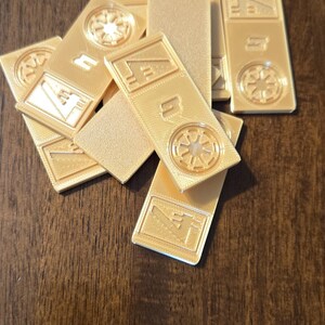 Może przedstawiać: Kolekcja złotych, prostokątnych żeton&oacute;w z wytłoczonymi wzorami. Niekt&oacute;re żetony mają okrągły emblemat, a inne kształty geometryczne. Żetony są rozrzucone na ciemnej drewnianej powierzchni.