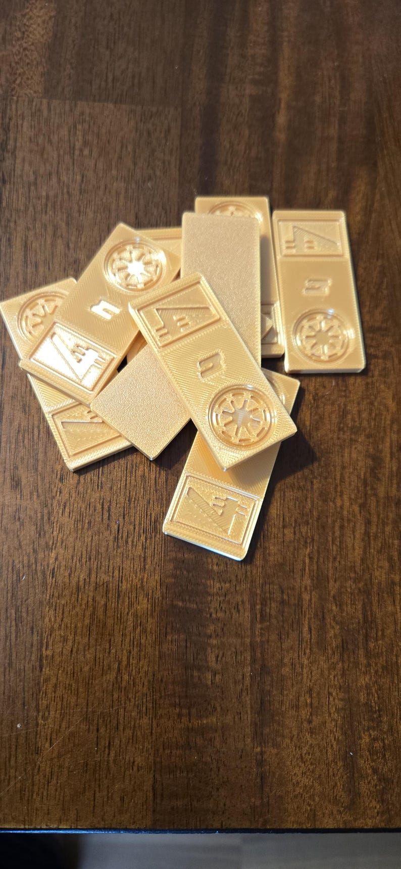 Może przedstawiać: Kolekcja złotych, prostokątnych żeton&oacute;w z wytłoczonymi wzorami. Niekt&oacute;re żetony mają okrągły emblemat, a inne kształty geometryczne. Żetony są rozrzucone na ciemnej drewnianej powierzchni.