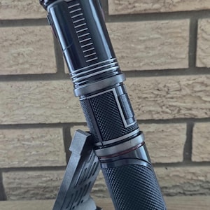 Op de afbeelding: Een zwart lightsaber met een rode accent en een textuur grip. De lightsaber staat op een zwarte standaard.