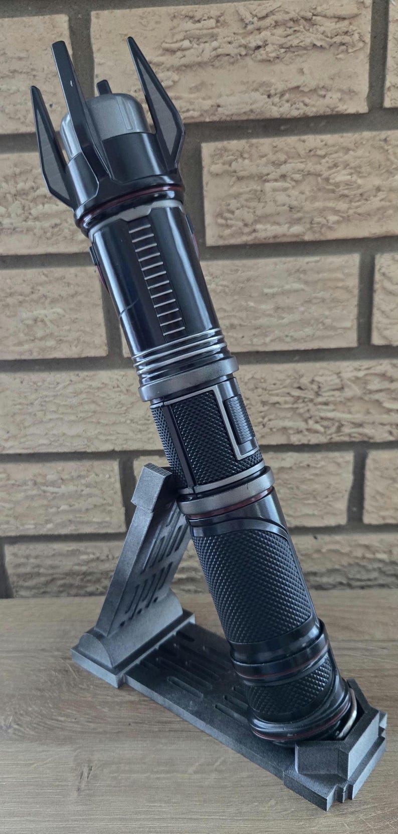 Op de afbeelding: Een zwart lightsaber met een rode accent en een textuur grip. De lightsaber staat op een zwarte standaard.