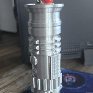 May include: A silver-colored lighter with a red button on top. The lighter is shaped like a cylinder with a rounded top and bottom. It has a detailed design with ridges and grooves.