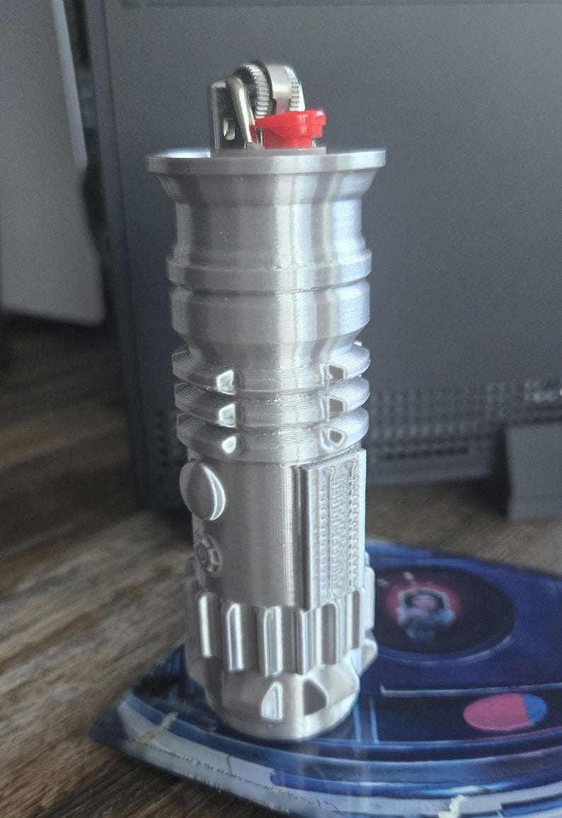 May include: A silver-colored lighter with a red button on top. The lighter is shaped like a cylinder with a rounded top and bottom. It has a detailed design with ridges and grooves.