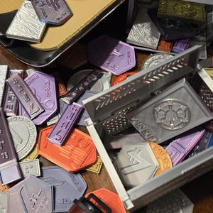 May include: A collection of small, colorful metal tokens with various designs and text. The tokens are scattered on a wooden surface, with some in a silver metal box. The box has a raised design of a star with a circle around it.