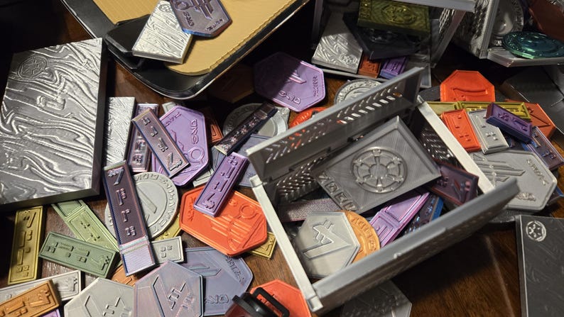 May include: A collection of small, colorful metal tokens with various designs and text. The tokens are scattered on a wooden surface, with some in a silver metal box. The box has a raised design of a star with a circle around it.