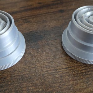 Puede incluir: Dos objetos de pl&aacute;stico gris con un dise&ntilde;o circular y base estriada se muestran sobre una superficie de madera. Los objetos tienen una apariencia en capas con un componente central detallado. Son componentes de un producto m&aacute;s grande.