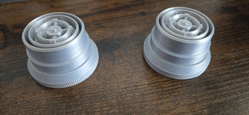 Puede incluir: Dos objetos de pl&aacute;stico gris con un dise&ntilde;o circular y base estriada se muestran sobre una superficie de madera. Los objetos tienen una apariencia en capas con un componente central detallado. Son componentes de un producto m&aacute;s grande.