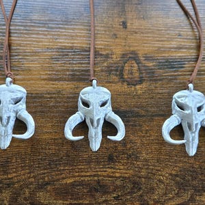 May include: Five identical pendants featuring a light gray, weathered skull-like creature design with curved horns, each suspended from a thin brown leather cord. The pendants exhibit a textured, matte finish with subtle dark shading in the crevices.