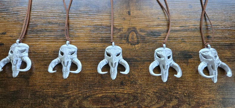 May include: Five identical pendants featuring a light gray, weathered skull-like creature design with curved horns, each suspended from a thin brown leather cord. The pendants exhibit a textured, matte finish with subtle dark shading in the crevices.