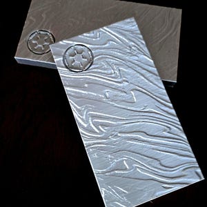 May include: Two grey textured rectangular tiles with a raised embossed symbol of a circle with a cross inside. The symbol is repeated on both tiles.