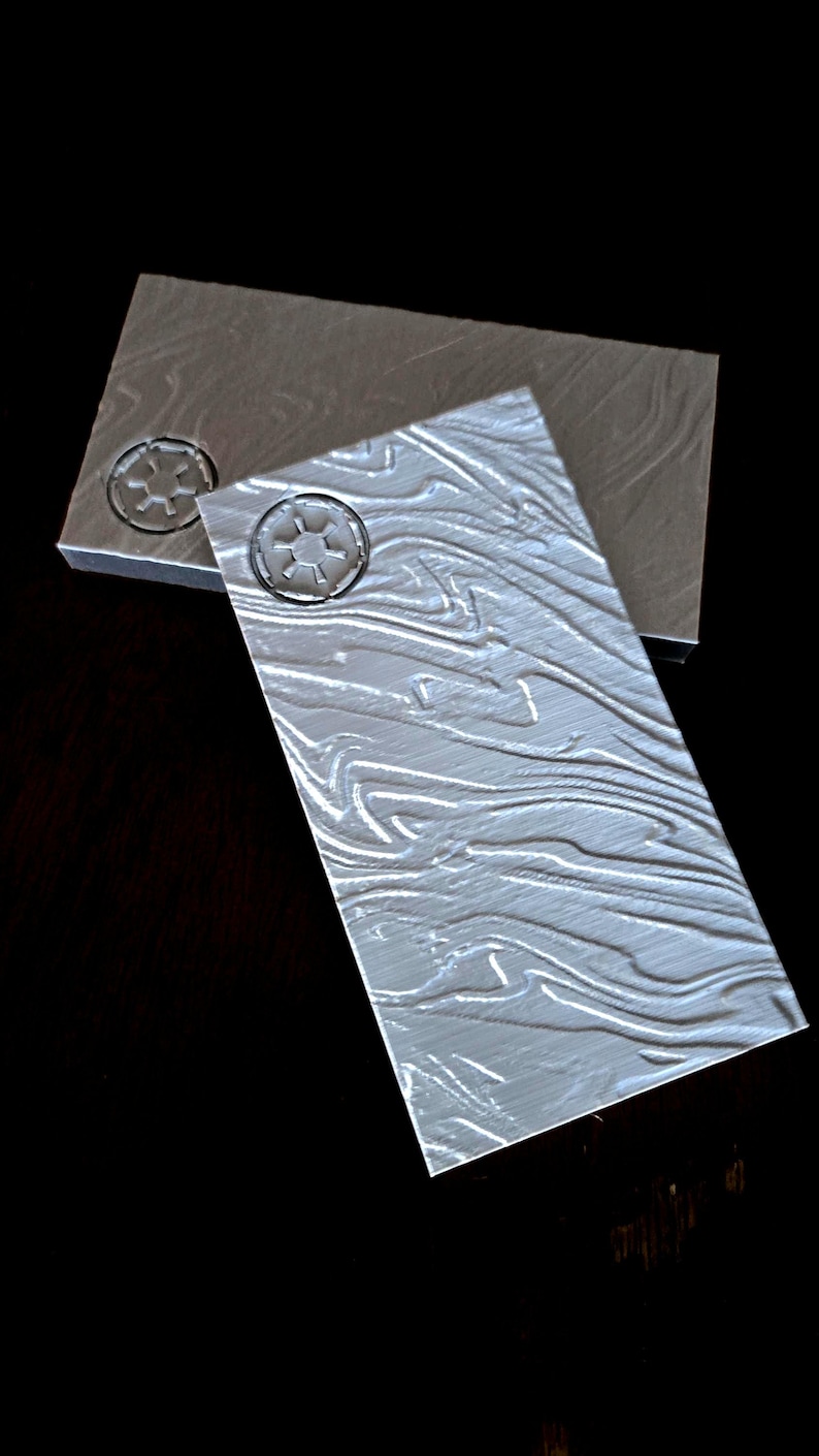 May include: Two grey textured rectangular tiles with a raised embossed symbol of a circle with a cross inside. The symbol is repeated on both tiles.