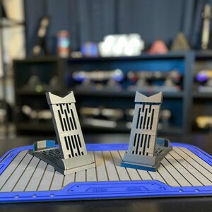 Peut inclure: Deux supports de t&eacute;l&eacute;phone gris et bleu au design unique, pos&eacute;s sur un tapis gris et bleu. Les supports pr&eacute;sentent un motif vertical &agrave; fentes et sont l&eacute;g&egrave;rement inclin&eacute;s. L'arri&egrave;re-plan montre des &eacute;tag&egrave;res avec divers objets.