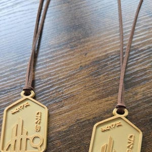 Puede incluir: Dos collares con colgantes dorados con cordones marrones. Cada colgante tiene forma hexagonal con un dise&ntilde;o arquitect&oacute;nico estilizado y texto. Los colgantes se muestran sobre una superficie de madera.
