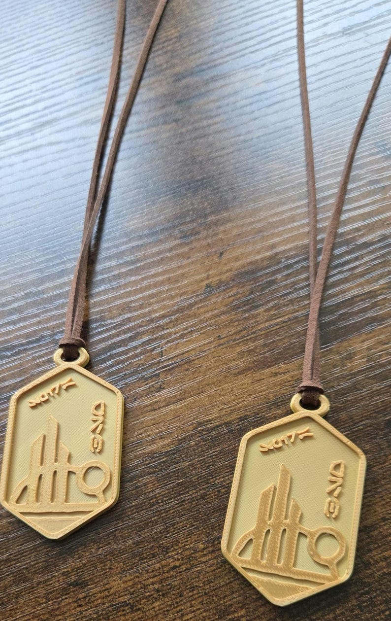 Puede incluir: Dos collares con colgantes dorados con cordones marrones. Cada colgante tiene forma hexagonal con un dise&ntilde;o arquitect&oacute;nico estilizado y texto. Los colgantes se muestran sobre una superficie de madera.