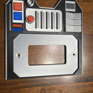 May include: A 3D printed gray and black panel with a rectangular cutout, a red button, and a row of five vertical lines. The panel has a black border and two small holes on the bottom.