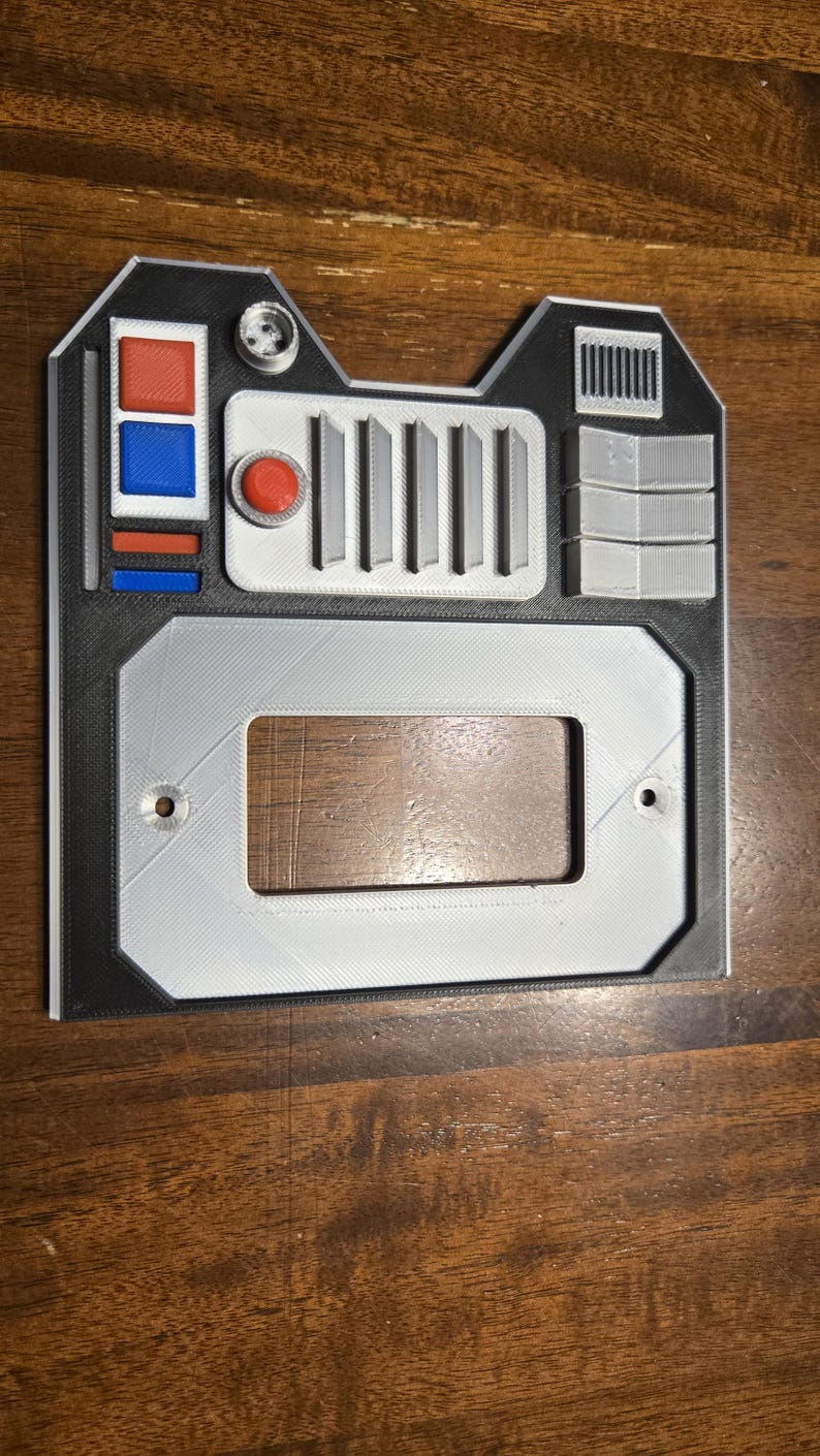 May include: A 3D printed gray and black panel with a rectangular cutout, a red button, and a row of five vertical lines. The panel has a black border and two small holes on the bottom.