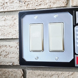May include: A black light switch cover with a silver panel and two toggle switches. The cover has a futuristic design with a silver button and two red and blue buttons.