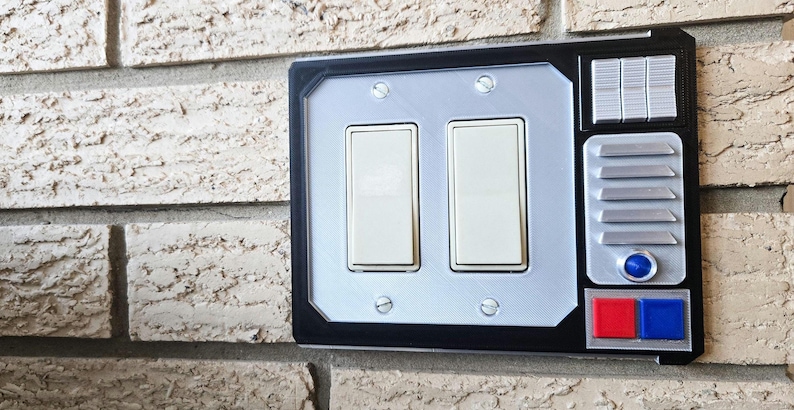 May include: A black light switch cover with a silver panel and two toggle switches. The cover has a futuristic design with a silver button and two red and blue buttons.