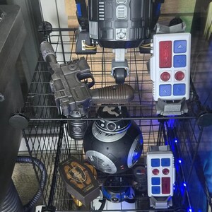 Peut inclure: Une collection de jouets et d'objets de collection sur le th&egrave;me de Star Wars, expos&eacute;e sur des &eacute;tag&egrave;res grillag&eacute;es. Comprend une grande figurine R2-D2 noire et argent&eacute;e, des dro&iuml;des BB-8, des blasters et des panneaux de contr&ocirc;le.