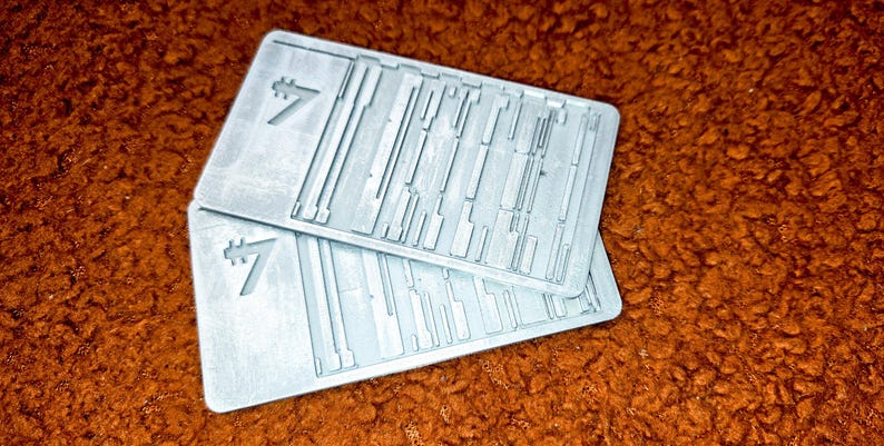 Peut inclure: Deux objets rectangulaires gris clair avec une surface textur&eacute;e et un chiffre sept stylis&eacute;. Les objets comportent une s&eacute;rie de lignes et de formes parall&egrave;les et perpendiculaires. Les objets sont empil&eacute;s l'un sur l'autre.