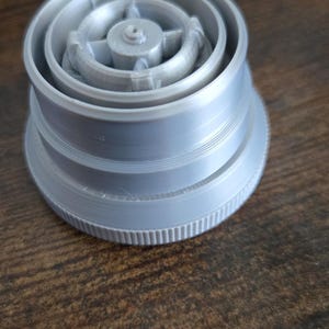 Puede incluir: Un objeto gris claro impreso en 3D con una base circular y m&uacute;ltiples anillos conc&eacute;ntricos. La parte superior presenta un cubo central con radios. El objeto es probablemente un componente o una pieza.
