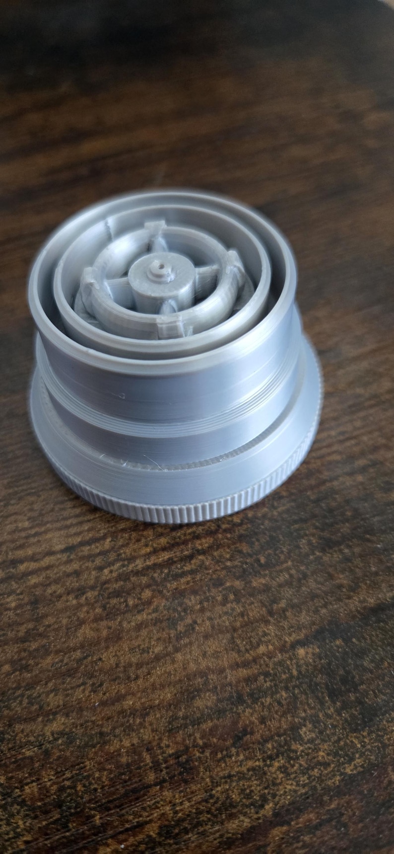 Puede incluir: Un objeto gris claro impreso en 3D con una base circular y m&uacute;ltiples anillos conc&eacute;ntricos. La parte superior presenta un cubo central con radios. El objeto es probablemente un componente o una pieza.