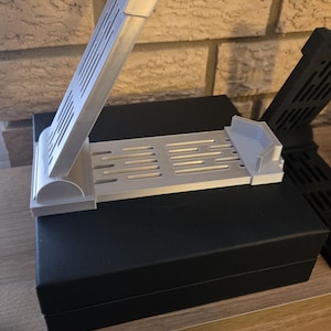 May include: A light gray 3D-printed phone stand with a slotted base and angled support. The stand is designed to hold a phone upright. Another black stand is visible in the background. The stand is sitting on a black box.