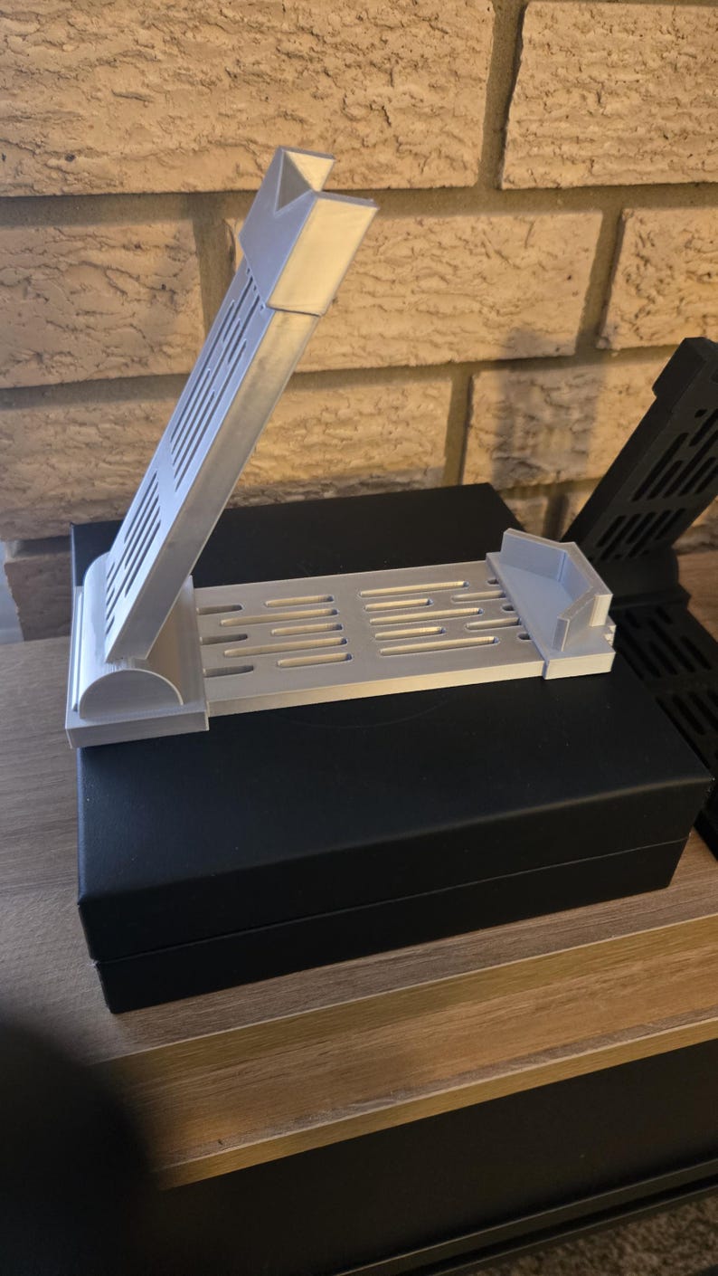 May include: A light gray 3D-printed phone stand with a slotted base and angled support. The stand is designed to hold a phone upright. Another black stand is visible in the background. The stand is sitting on a black box.