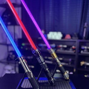 May include: Three illuminated lightsabers with blue, red, and purple blades are displayed on a black stand with a blue border. The hilts have metallic and black finishes. The background is dark with shelves.