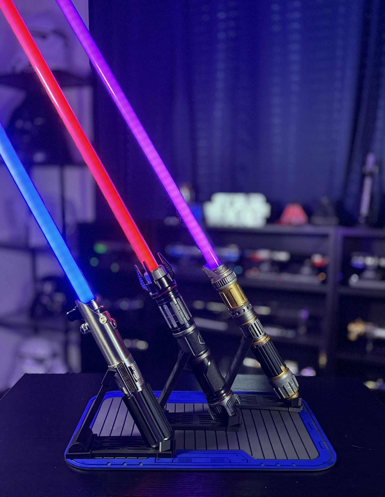 May include: Three illuminated lightsabers with blue, red, and purple blades are displayed on a black stand with a blue border. The hilts have metallic and black finishes. The background is dark with shelves.